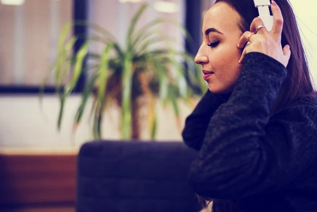 A relaxed woman with dark hair wearing a dark jumper and headphones, sitting in her home with a plant in the background. She appears calm and focused during an RTT session.