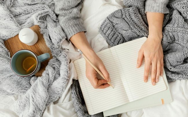 Young woman journalling in bed with a notebook and pen, creating a relaxing writing environment