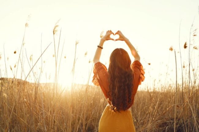 A woman standing in a sunlit field, forming a heart shape with her hands, symbolising love, self-acceptance, and connection.
