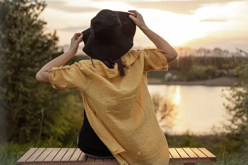 Woman standing outdoors holding onto her hat while looking out across the water