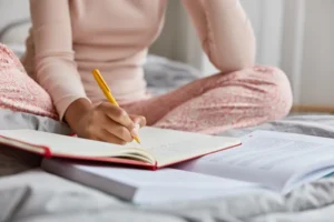 Woman sitting on a bed writing thoughtfully in a notebook in soft pink pyjamas