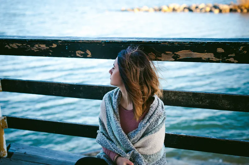 Woman wrapped in a blue shawl sitting by the sea and looking out across the water