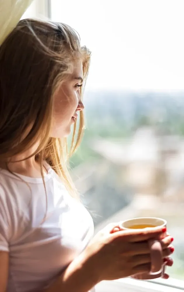 A woman sitting by a window with a cup, reflecting in a calm moment that represents the supportive space offered through online counselling at Clear Haven Therapy.