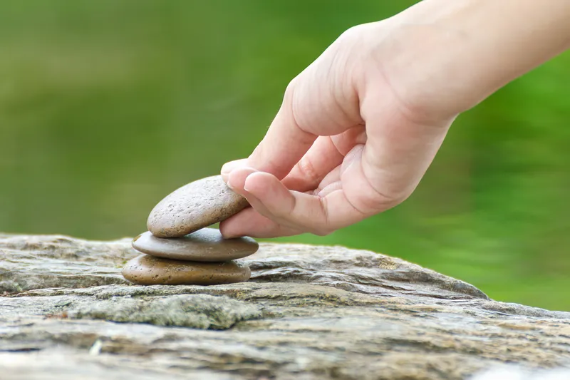 Hand carefully stacking smooth stones on a rock beside still green water