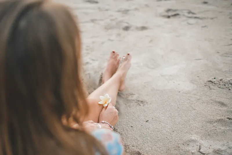 Woman sitting on a sandy beach holding a small white flower by her feet