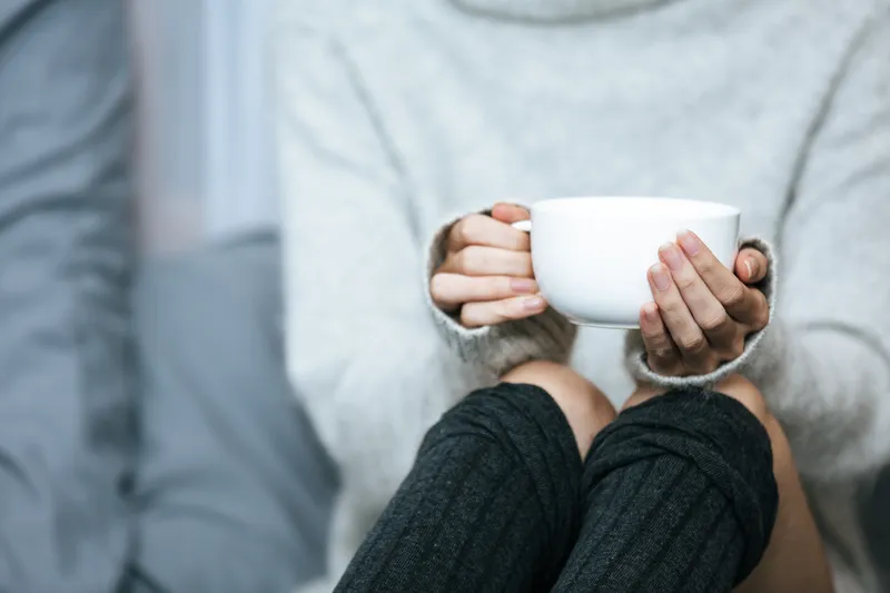 Close up of hands wrapped cosily around a white mug with knitted sleeves in soft light