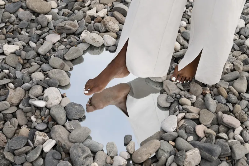Bare feet standing on smooth pebbles beside a still puddle reflecting the sky
