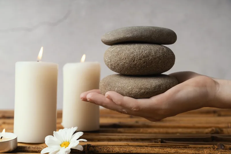 Hand holding three smooth stacked stones beside lit candles and a white flower