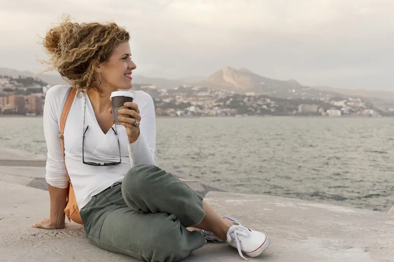 Woman sitting by the sea holding a warm drink during a quiet moment of reflection