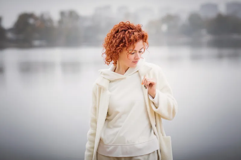 Woman standing outdoors in a white coat, looking down thoughtfully